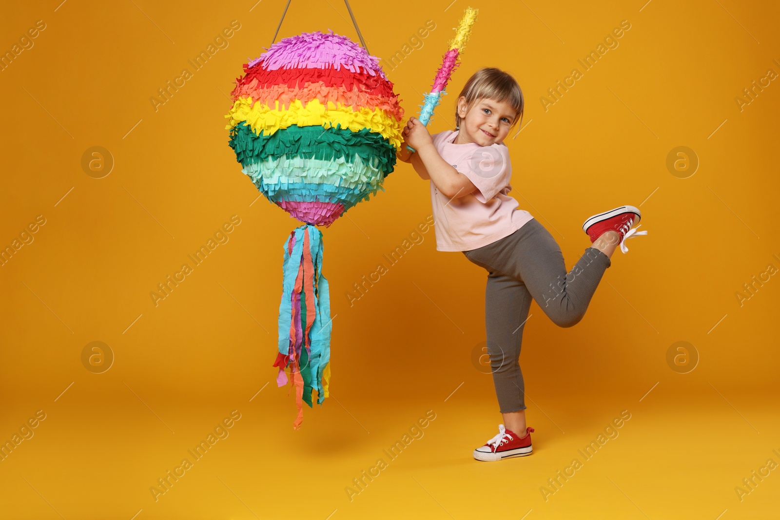 Photo of Cute girl breaking bright pinata with stick on orange background