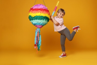 Photo of Cute girl breaking bright pinata with stick on orange background