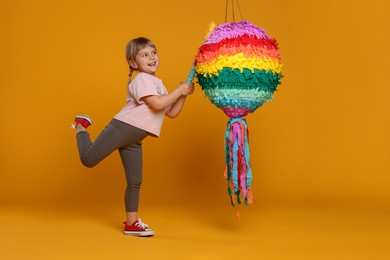 Photo of Cute girl breaking bright pinata with stick on orange background