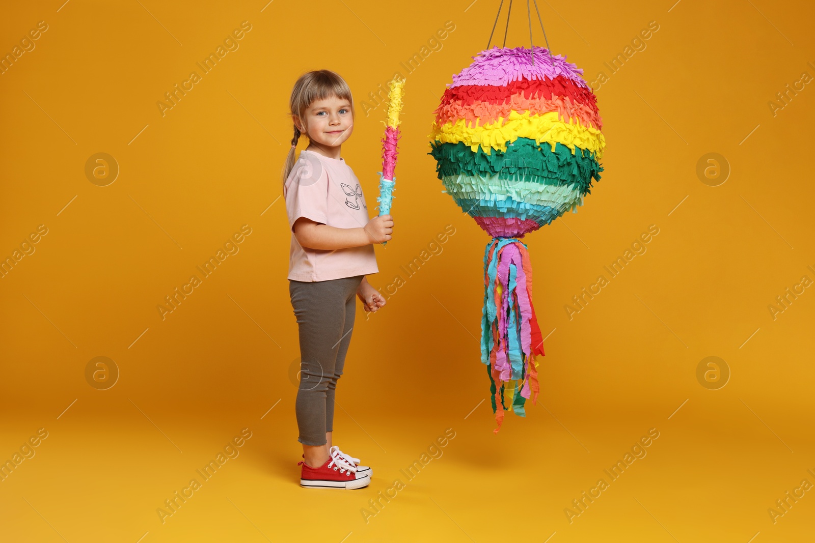 Photo of Cute girl with bright pinata and stick on orange background