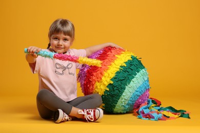 Photo of Cute girl with bright pinata and stick on orange background