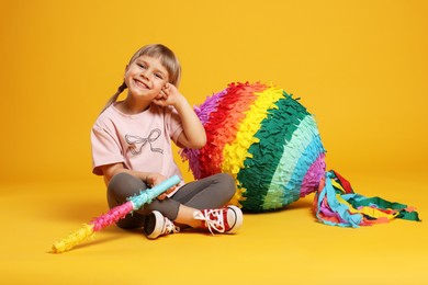 Happy girl with bright pinata and stick on orange background