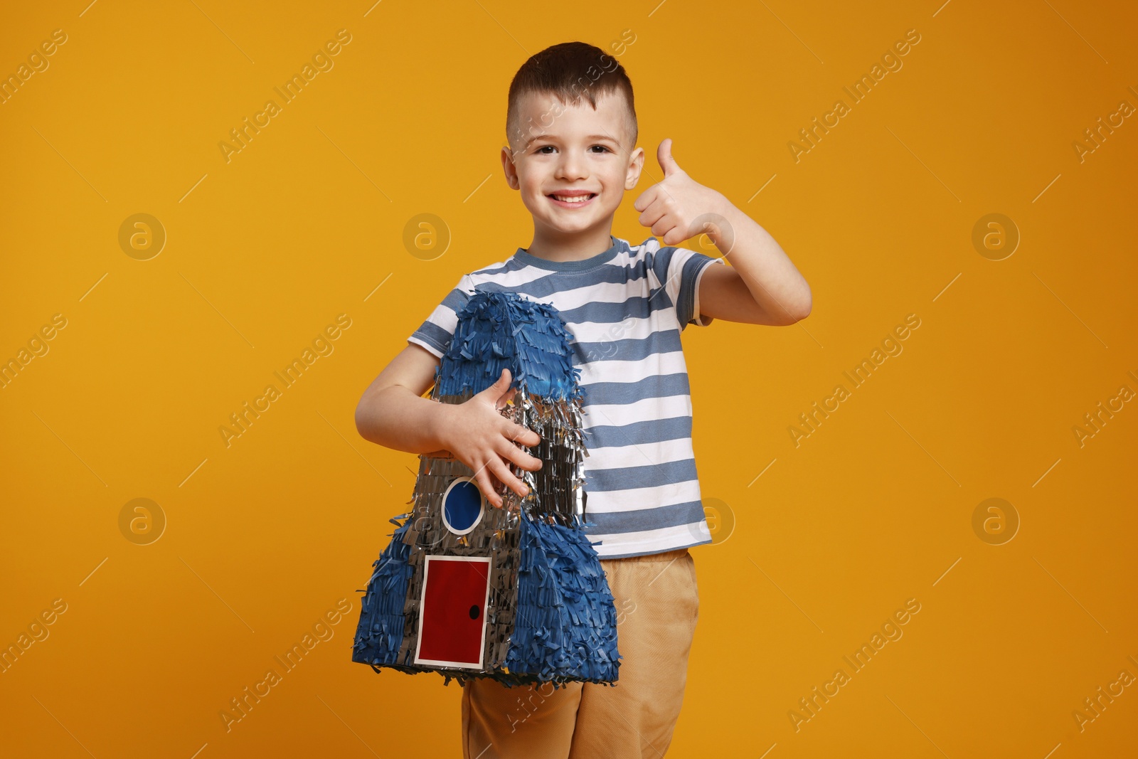 Photo of Happy boy with rocket shaped pinata showing thumbs up on orange background