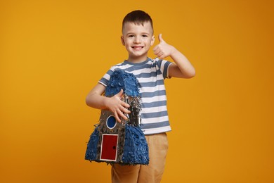 Photo of Happy boy with rocket shaped pinata showing thumbs up on orange background