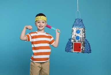 Happy boy with rocket shaped pinata and stick on light blue background