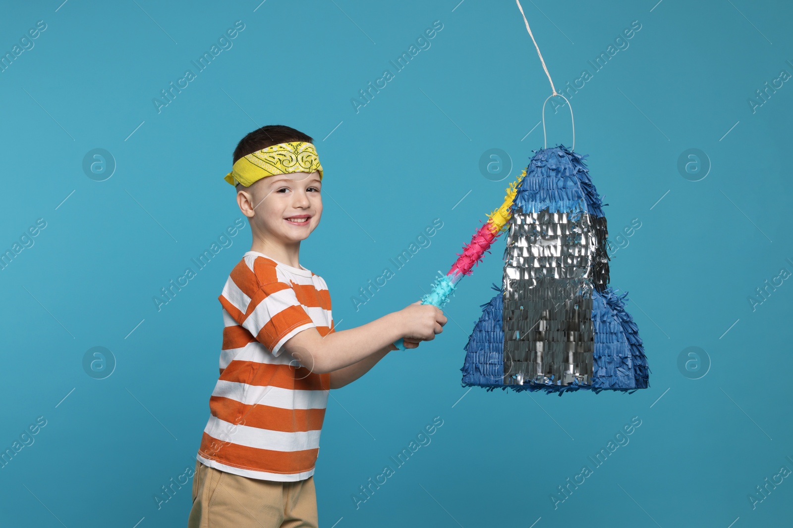 Photo of Happy boy breaking rocket shaped pinata with stick on light blue background