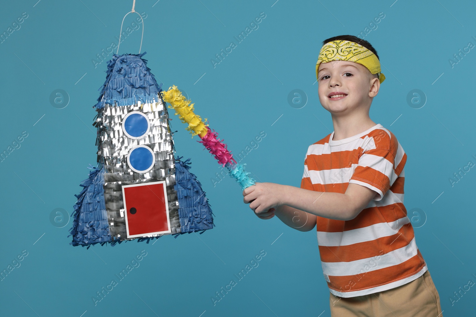 Photo of Happy boy breaking rocket shaped pinata with stick on light blue background