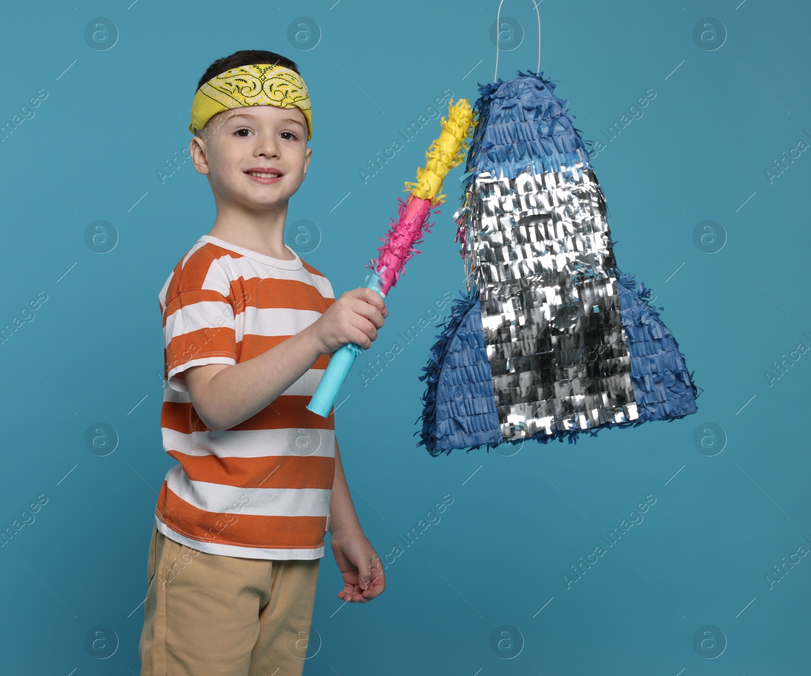 Photo of Happy boy breaking rocket shaped pinata with stick on light blue background