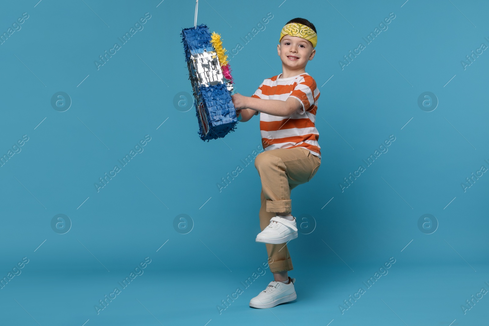 Photo of Happy boy breaking rocket shaped pinata with stick on light blue background