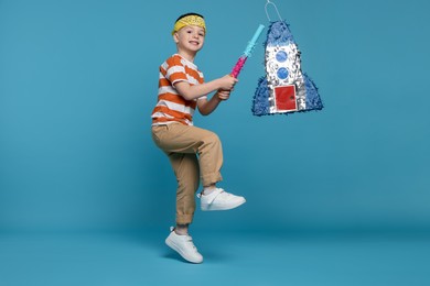 Photo of Happy boy breaking rocket shaped pinata with stick on light blue background