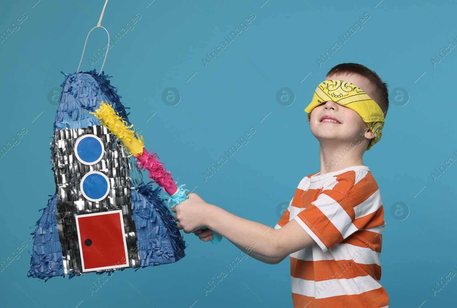 Photo of Happy boy with tied eyes breaking rocket shaped pinata on light blue background