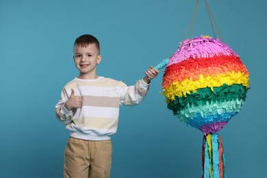 Happy boy with bright pinata and stick showing thumbs up on light blue background