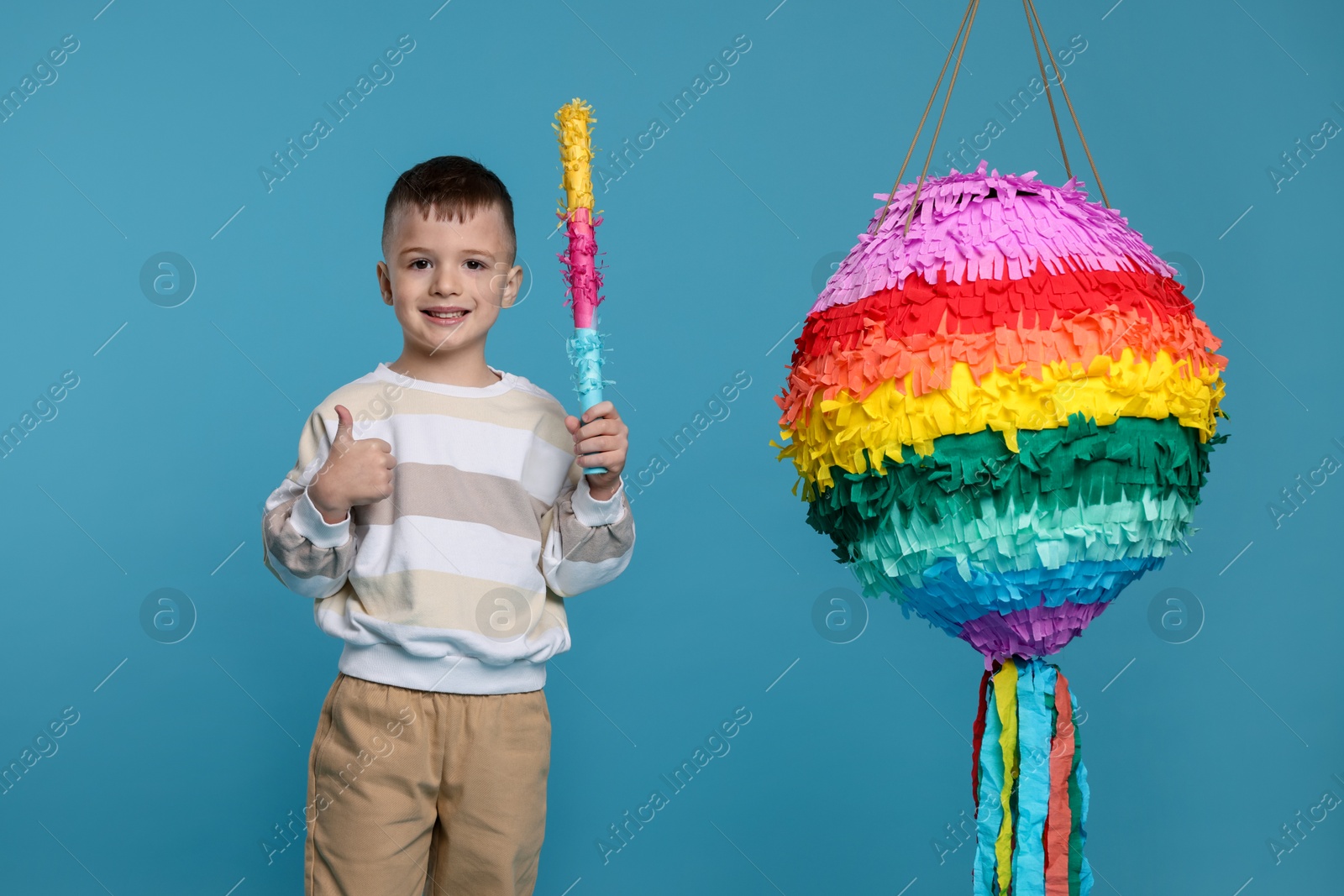 Photo of Happy boy with bright pinata and stick showing thumbs up on light blue background