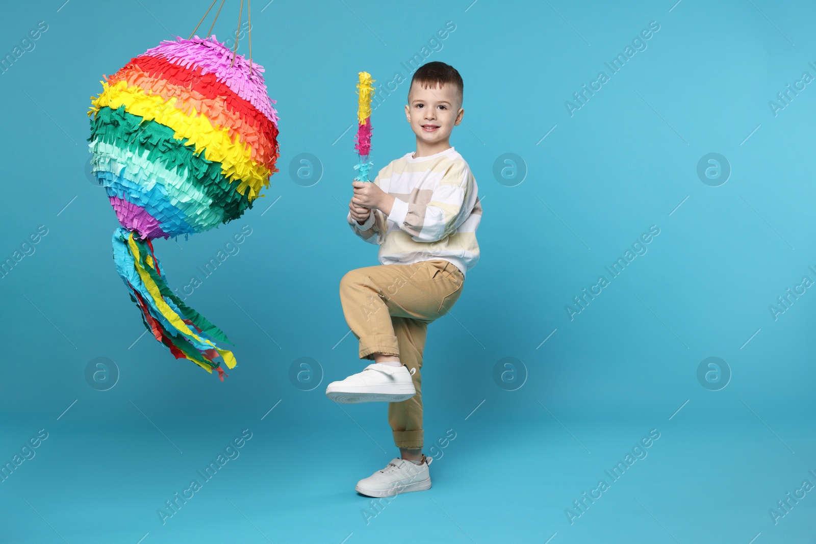 Photo of Happy boy breaking bright pinata with stick on light blue background