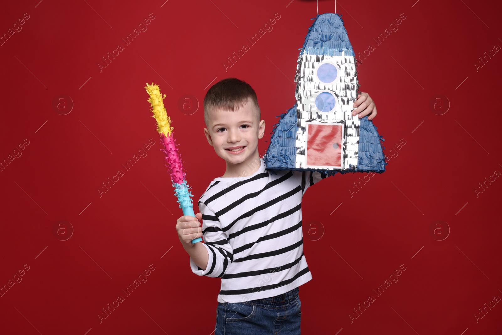 Photo of Happy boy with rocket shaped pinata and stick on red background