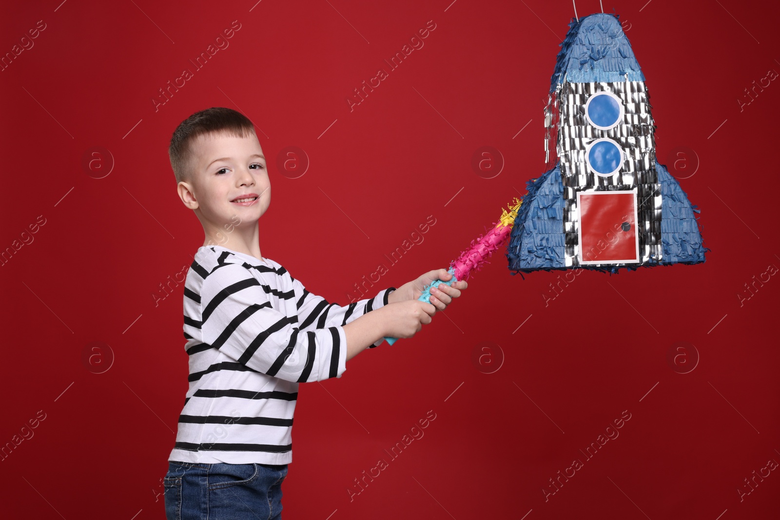 Photo of Happy boy breaking rocket shaped pinata with stick on red background