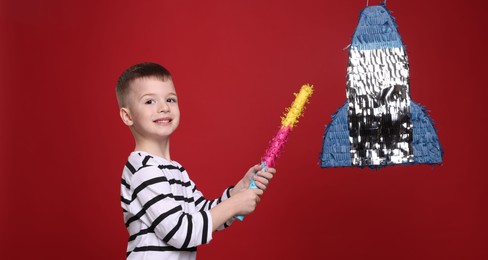 Happy boy breaking rocket shaped pinata with stick on red background