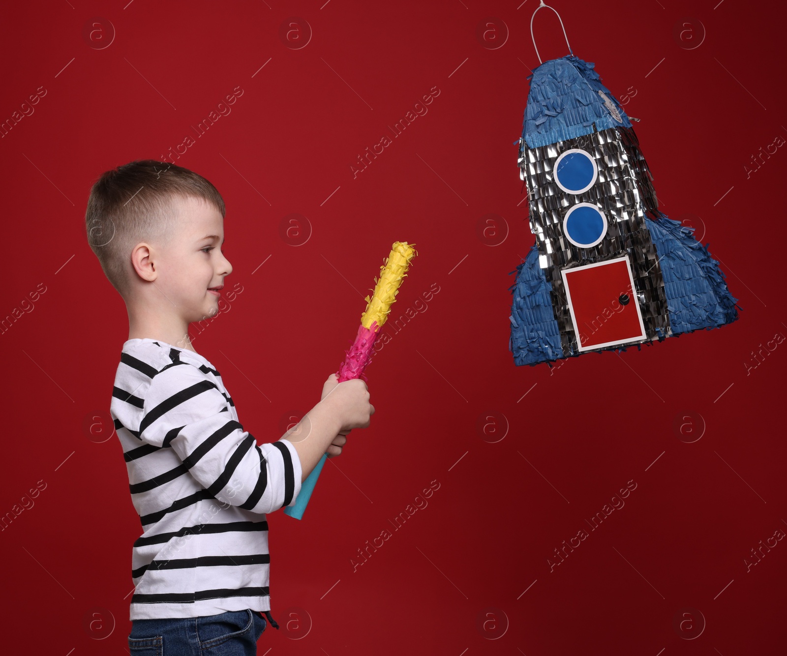 Photo of Happy boy breaking rocket shaped pinata with stick on red background