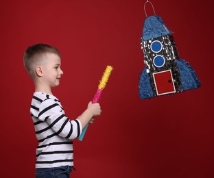 Photo of Happy boy breaking rocket shaped pinata with stick on red background