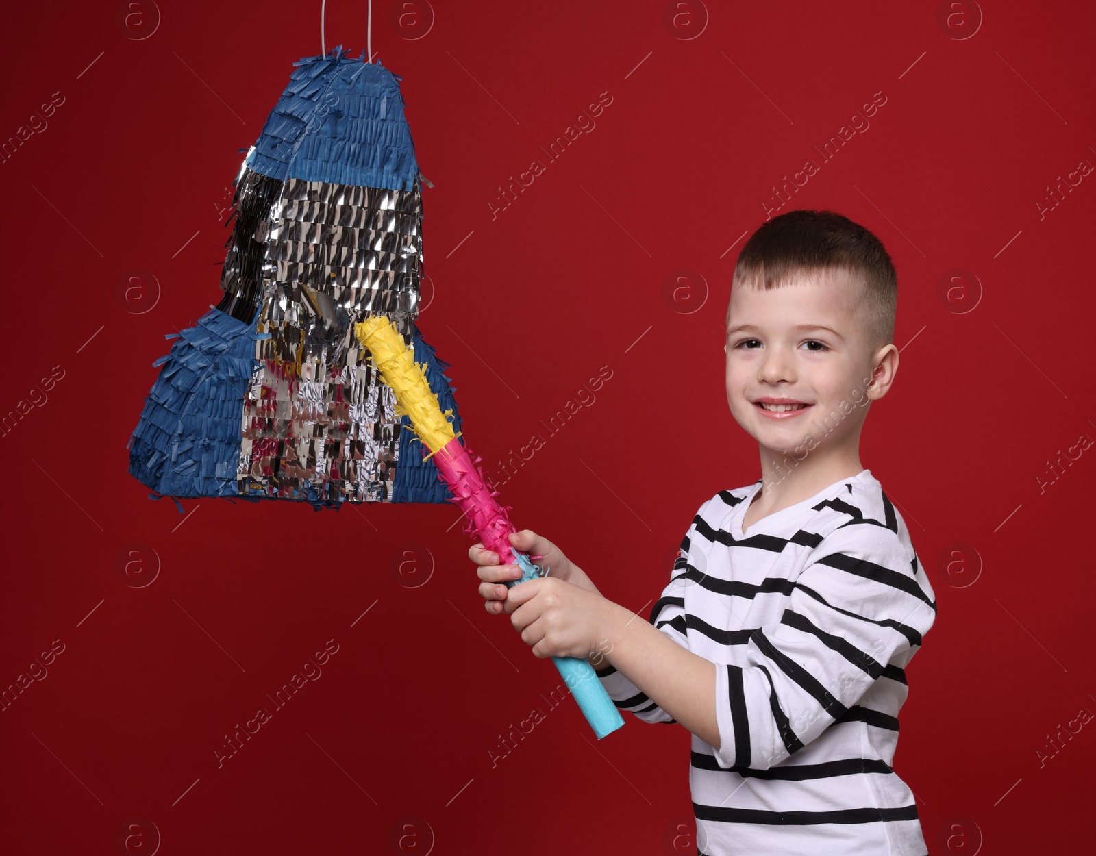 Photo of Happy boy breaking rocket shaped pinata with stick on red background