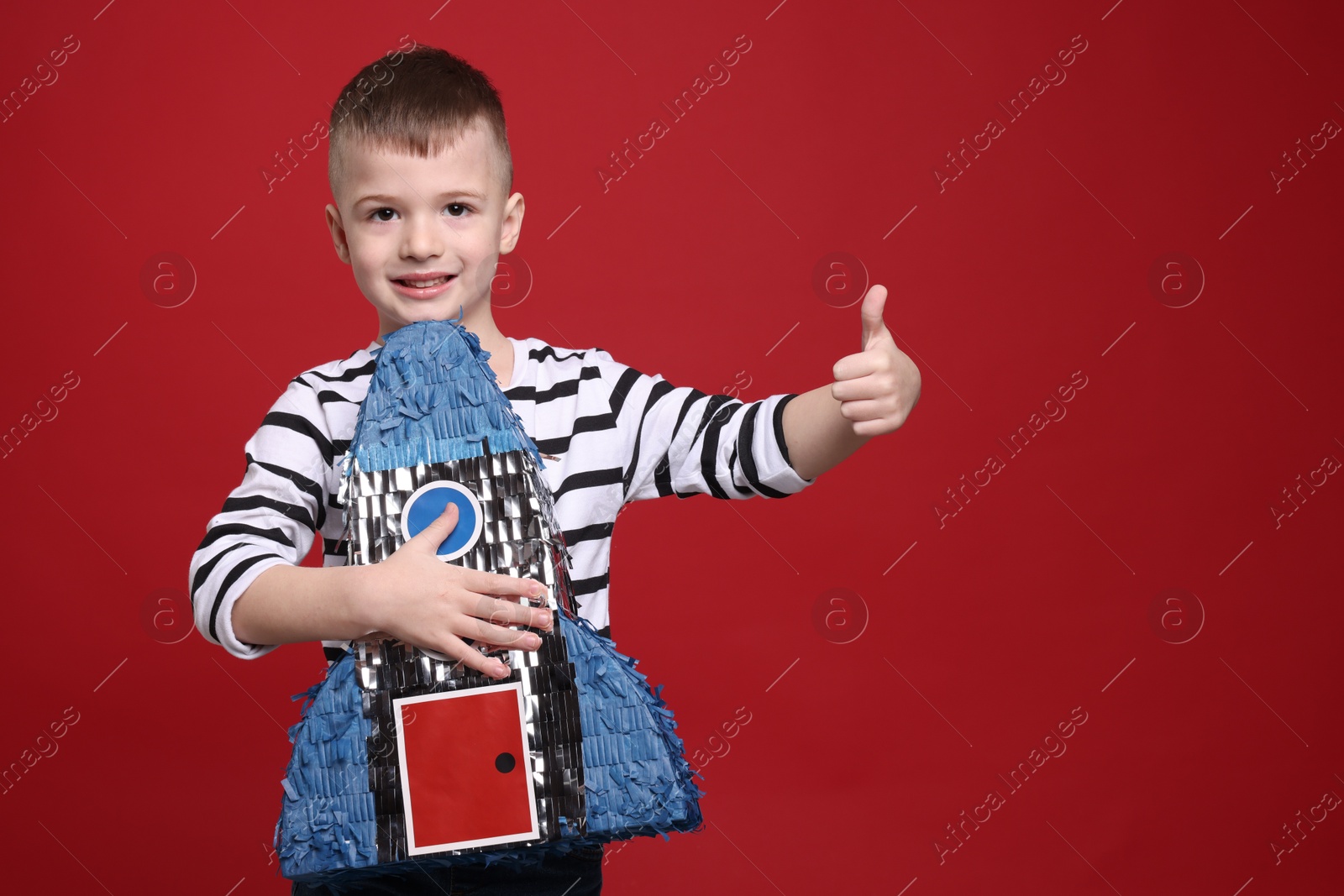 Photo of Happy boy with rocket shaped pinata showing thumbs up on red background. Space for text