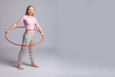 Beautiful young woman exercising with hula hoop on grey background, space for text