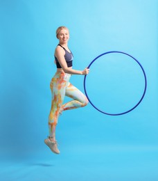 Beautiful young woman exercising with hula hoop on light blue background