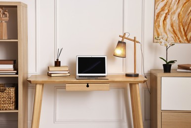 Photo of Comfortable workplace with modern laptop and lamp on wooden desk in home office