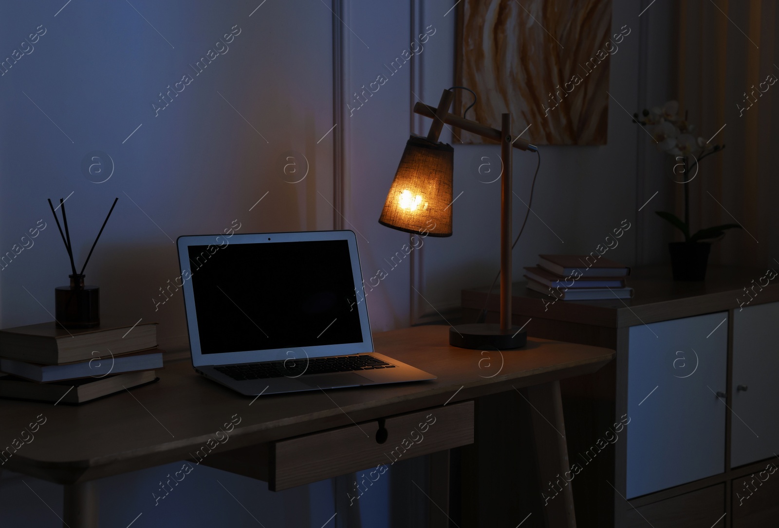 Photo of Comfortable workplace with modern laptop and lamp on wooden desk at home office in evening