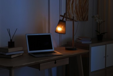 Photo of Comfortable workplace with modern laptop and lamp on wooden desk at home office in evening