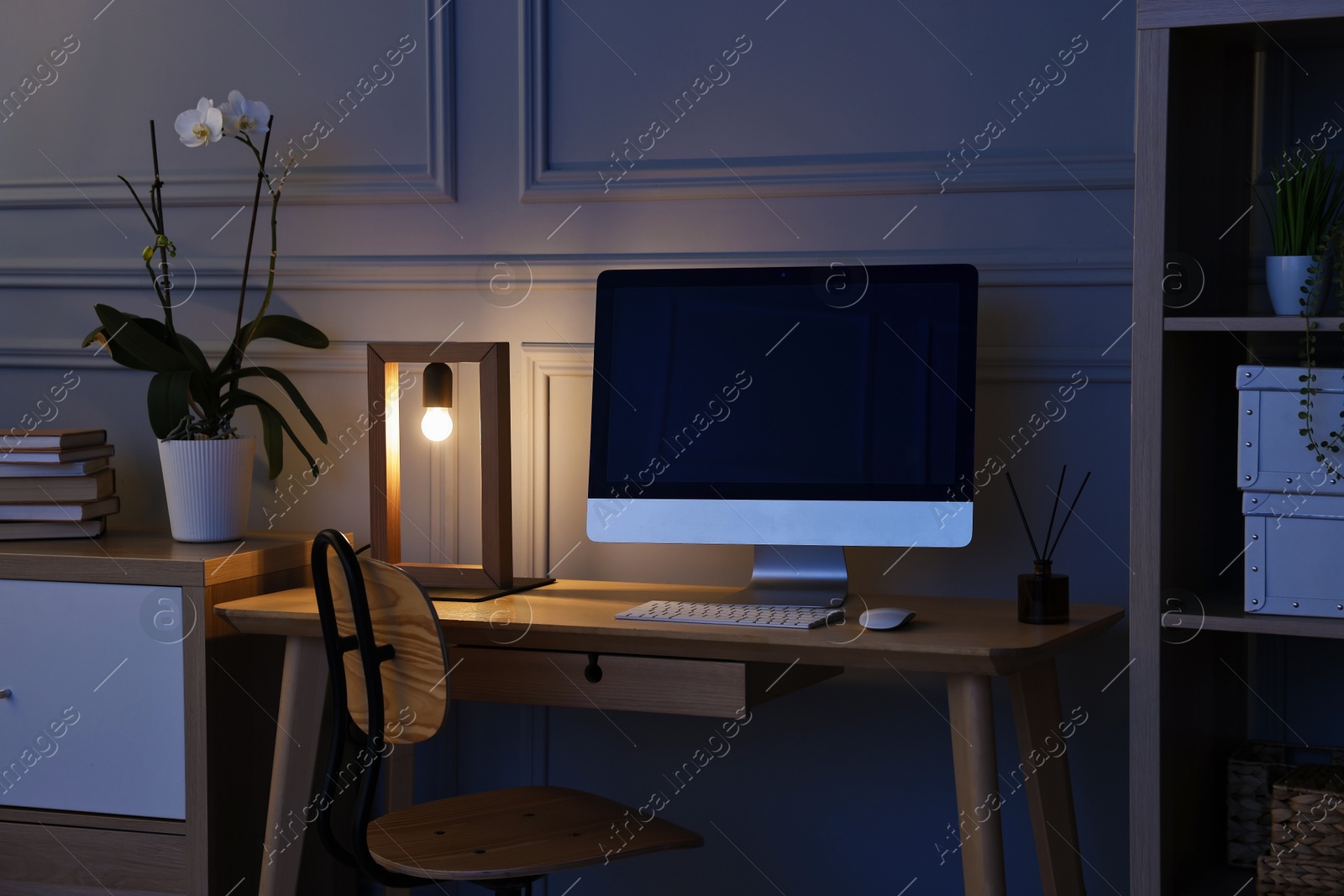 Photo of Comfortable workplace with modern computer and lamp on wooden desk at home office in evening