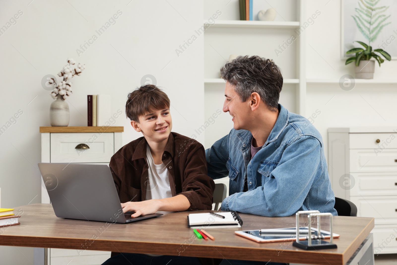 Photo of Smiling father and his son doing homework with laptop at table indoors