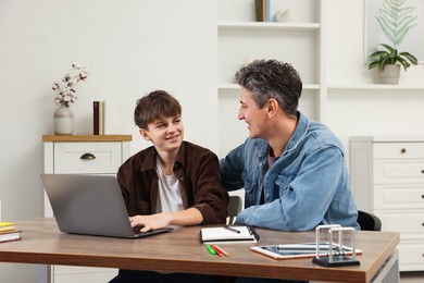 Smiling father and his son doing homework with laptop at table indoors