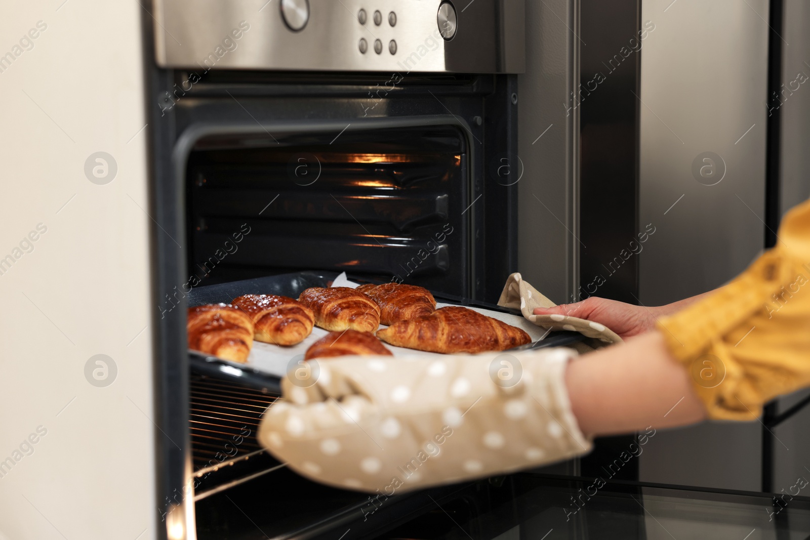 Photo of Woman taking baking sheet with tasty croissants out of oven indoors, closeup