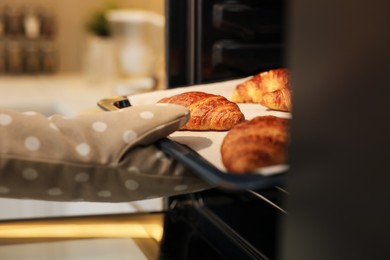 Woman taking baking sheet with tasty croissants out of oven indoors, closeup