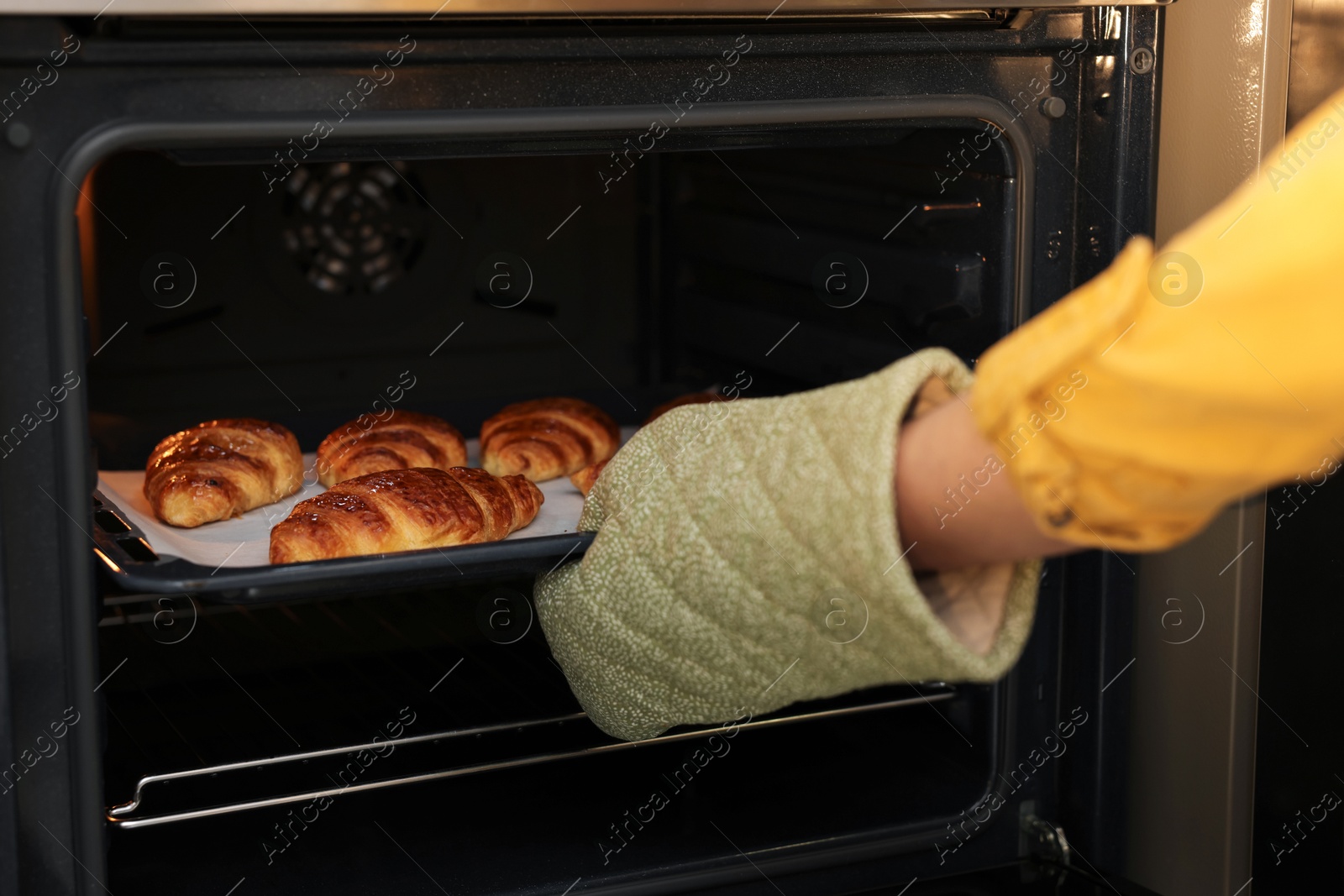 Photo of Woman taking baking sheet with tasty croissants out of oven, closeup