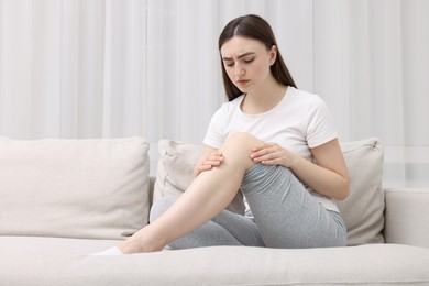 Young woman suffering from pain in knee on sofa at home. Space for text