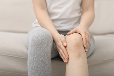 Young woman suffering from pain in knee on sofa at home, closeup