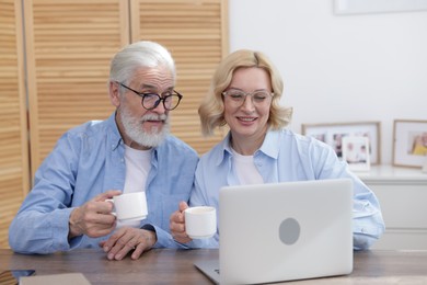 Senior man and mature woman with coffee watching something on laptop at home. Happy couple