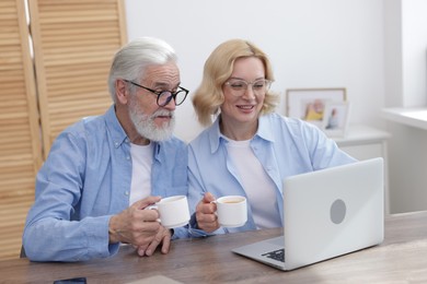 Senior man and mature woman with coffee watching something on laptop at home. Happy couple