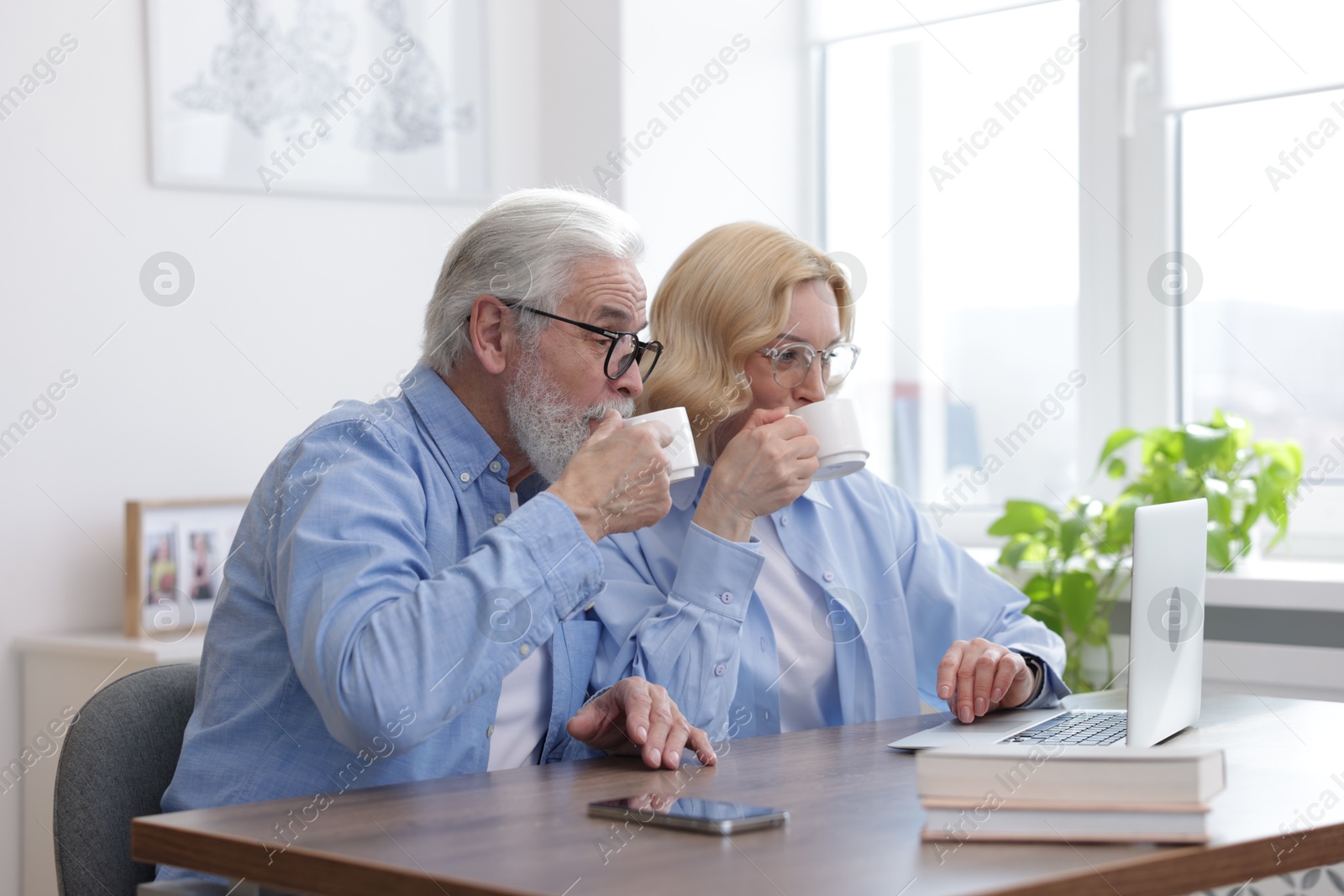 Photo of Senior man and mature woman drinking coffee while watching something on laptop at home. Happy couple