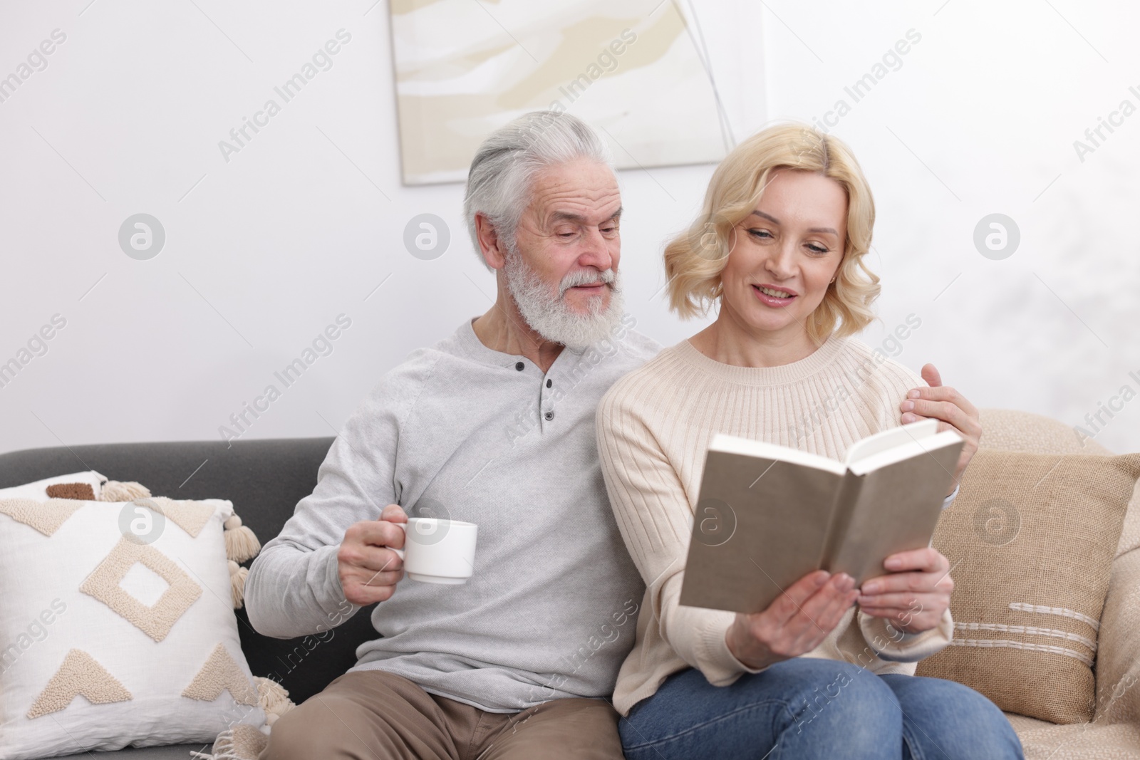 Photo of Senior man with coffee and mature woman reading book at home. Happy couple