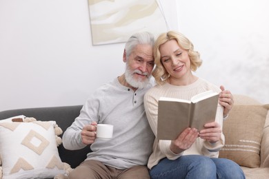 Photo of Senior man with coffee and mature woman reading book at home. Happy couple