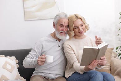 Photo of Senior man with coffee and mature woman reading book at home. Happy couple