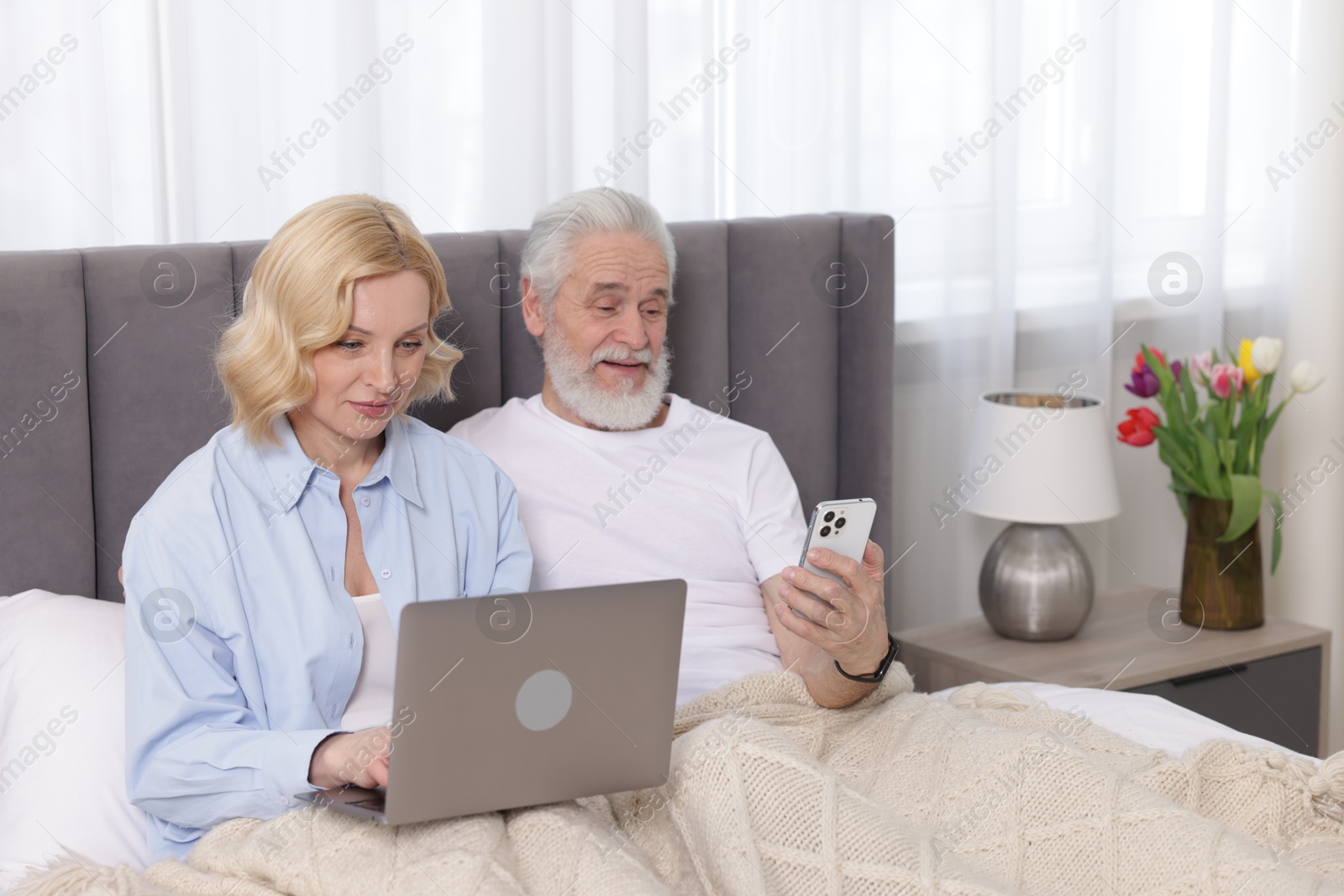 Photo of Senior man with smartphone and mature woman using laptop at home. Happy couple