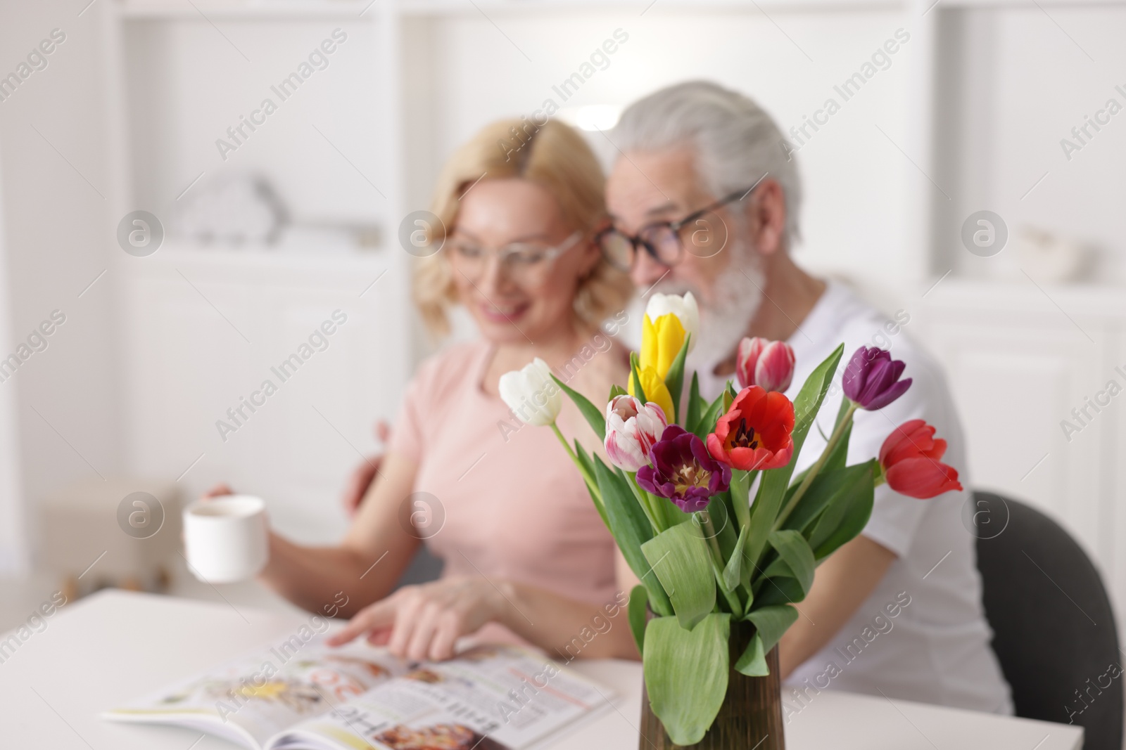 Photo of Senior man and mature woman spending time together at home, focus on tulips. Happy couple