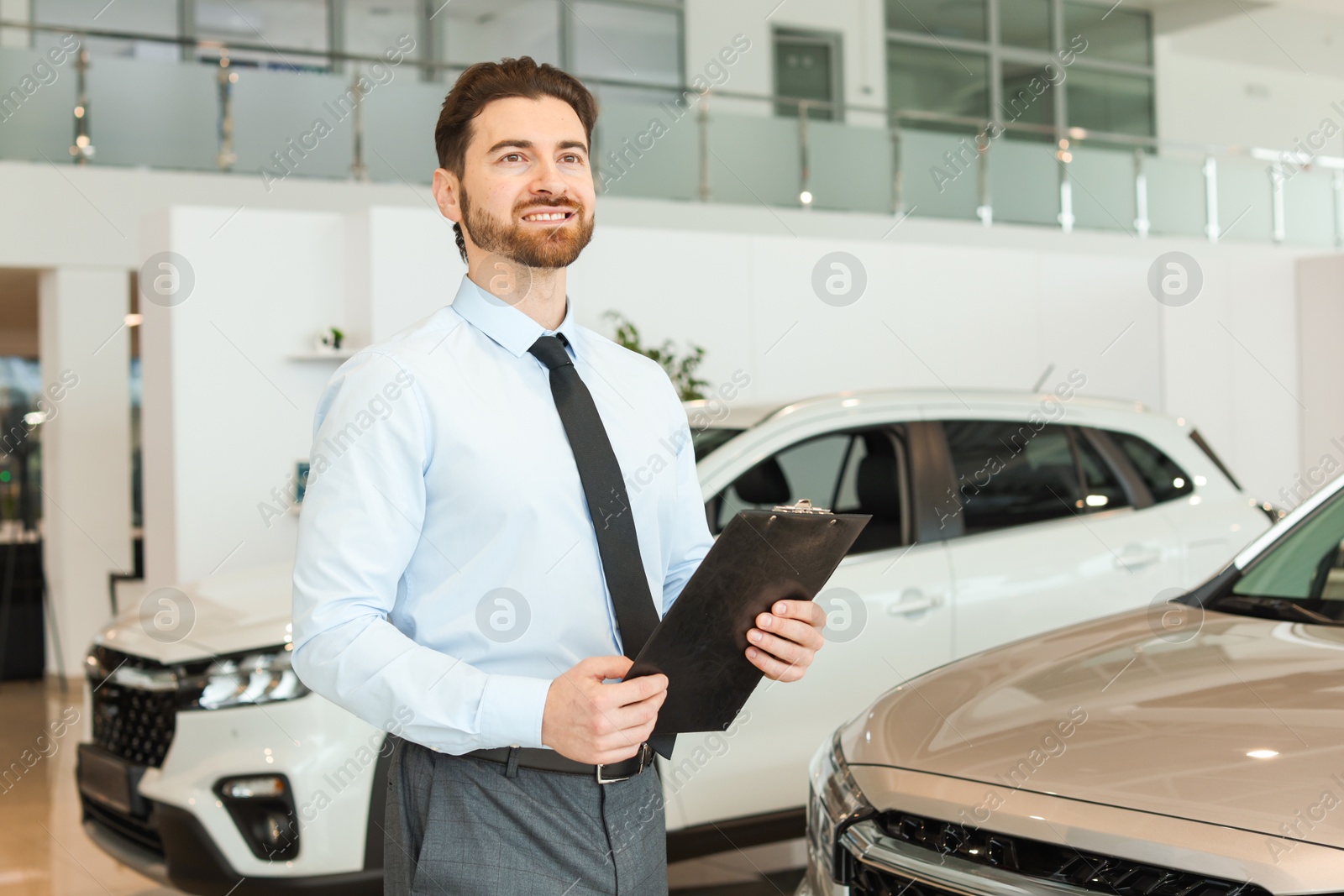 Photo of Happy salesman with clipboard near new cars in salon