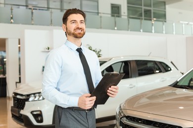 Happy salesman with clipboard near new cars in salon