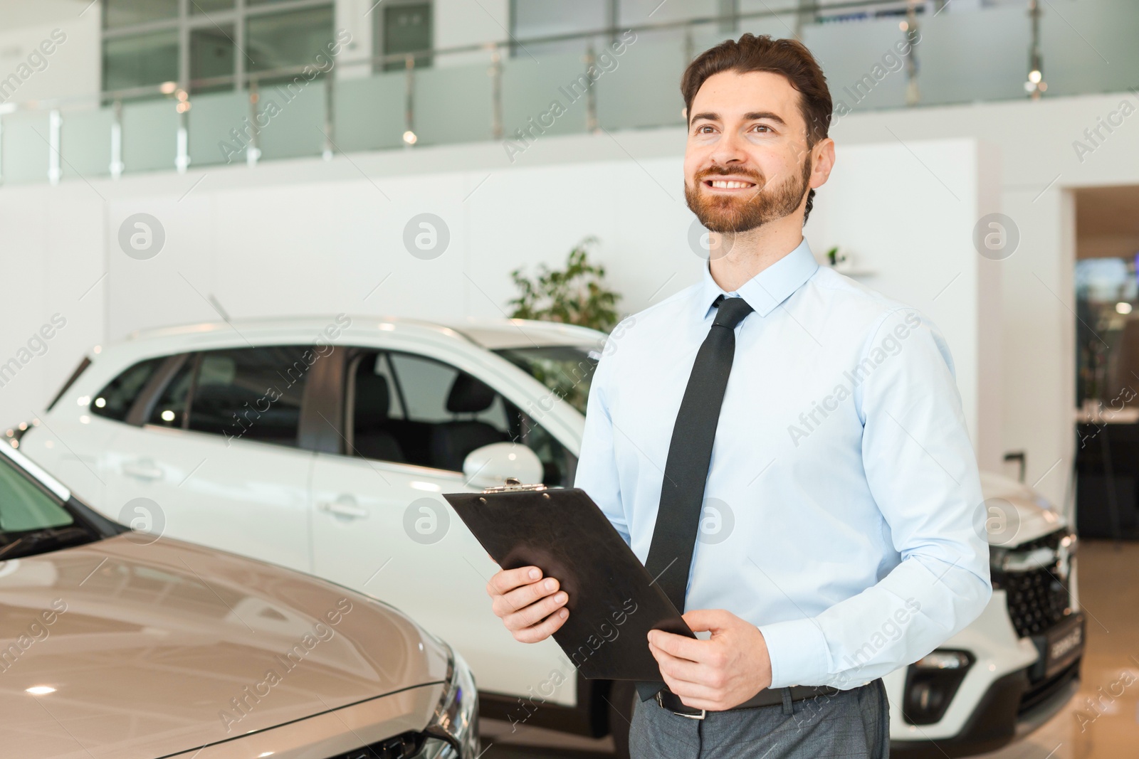 Photo of Happy salesman with clipboard near new cars in salon