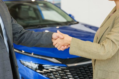 Photo of Saleswoman and client shaking hands near new car in salon, closeup